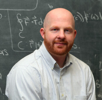 UIC Senior Lecturer, Andrew Shulman, smiling in blue checked shirt in front of chalkboard 