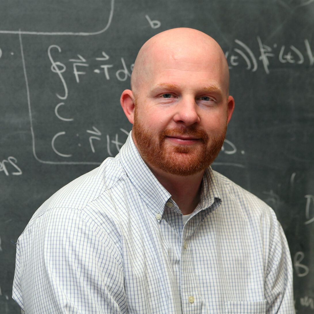 UIC Senior Lecturer, Andrew Shulman, smiling in blue checked shirt in front of chalkboard