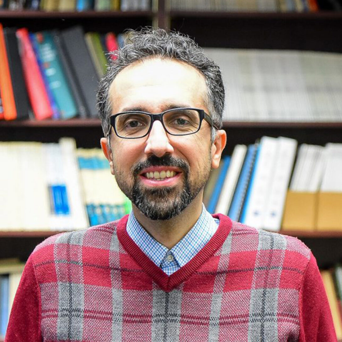 Dr. Hossein Ataei smiles at camera in red plaid shirt in front of bookshelf