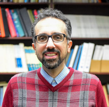 Dr. Hossein Ataei smiles at camera in red plaid shirt in front of bookshelf 